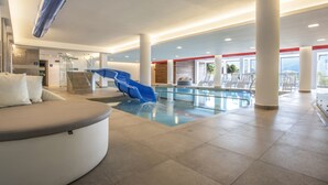 Un área de piscina interior moderna con un tobogán azul, una zona de estar con un banco curvo y vistas al exterior a través de amplios ventanales.
