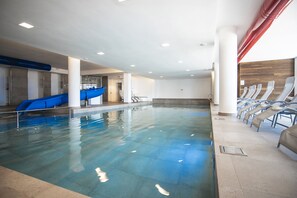 Una piscina interior con un tobogán azul, sillones de descanso y suelo de baldosas.