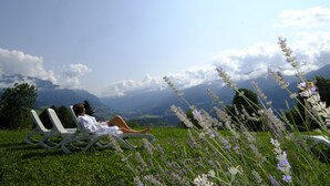 Una persona descansando en una silla reclinable blanca en un campo cubierto de hierba y flores de lavanda, con vistas a un paisaje montañoso.