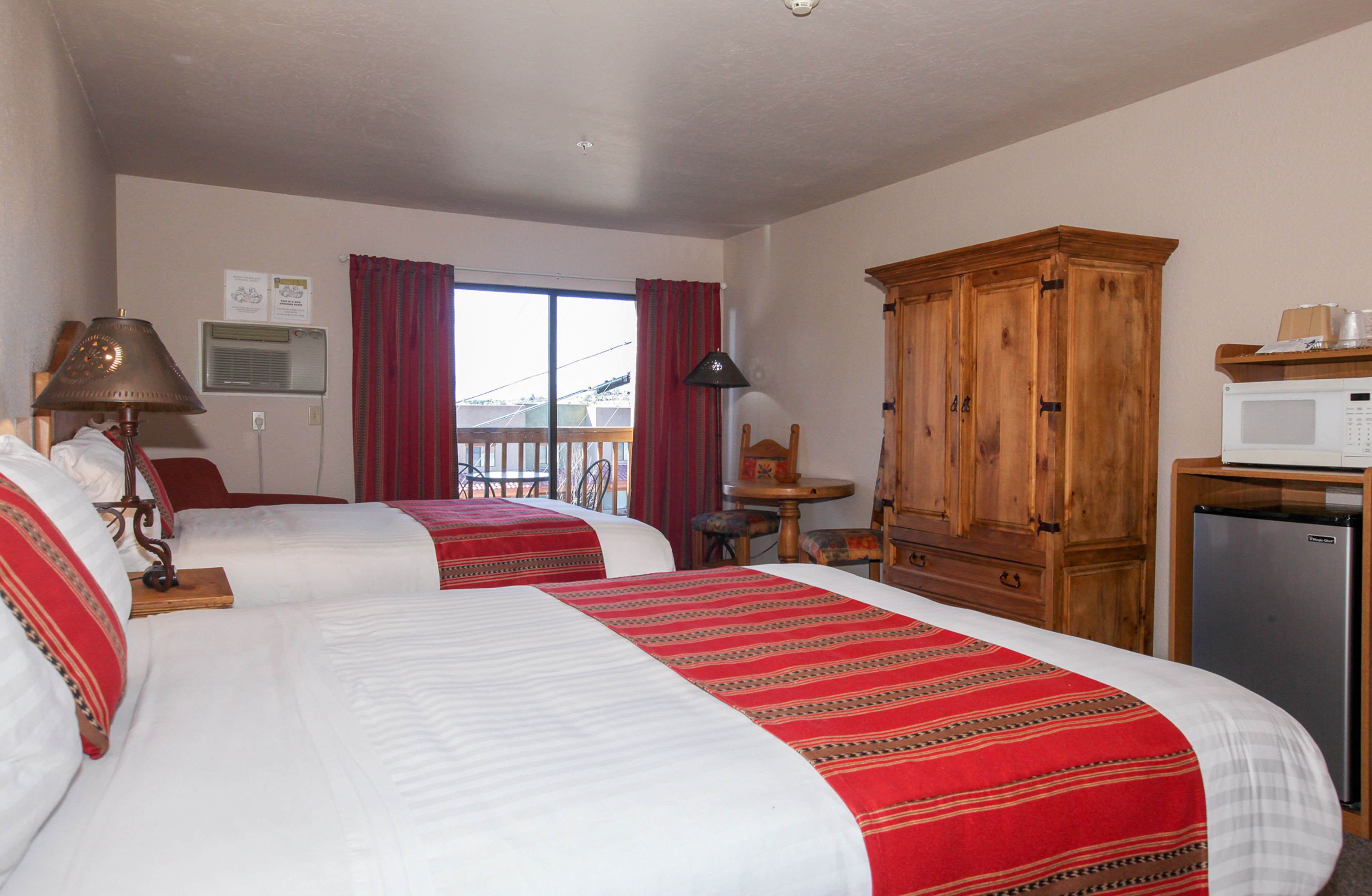 A hotel room with two beds, a wooden cabinet, a desk, a chair, a lamp, a microwave, and a refrigerator.