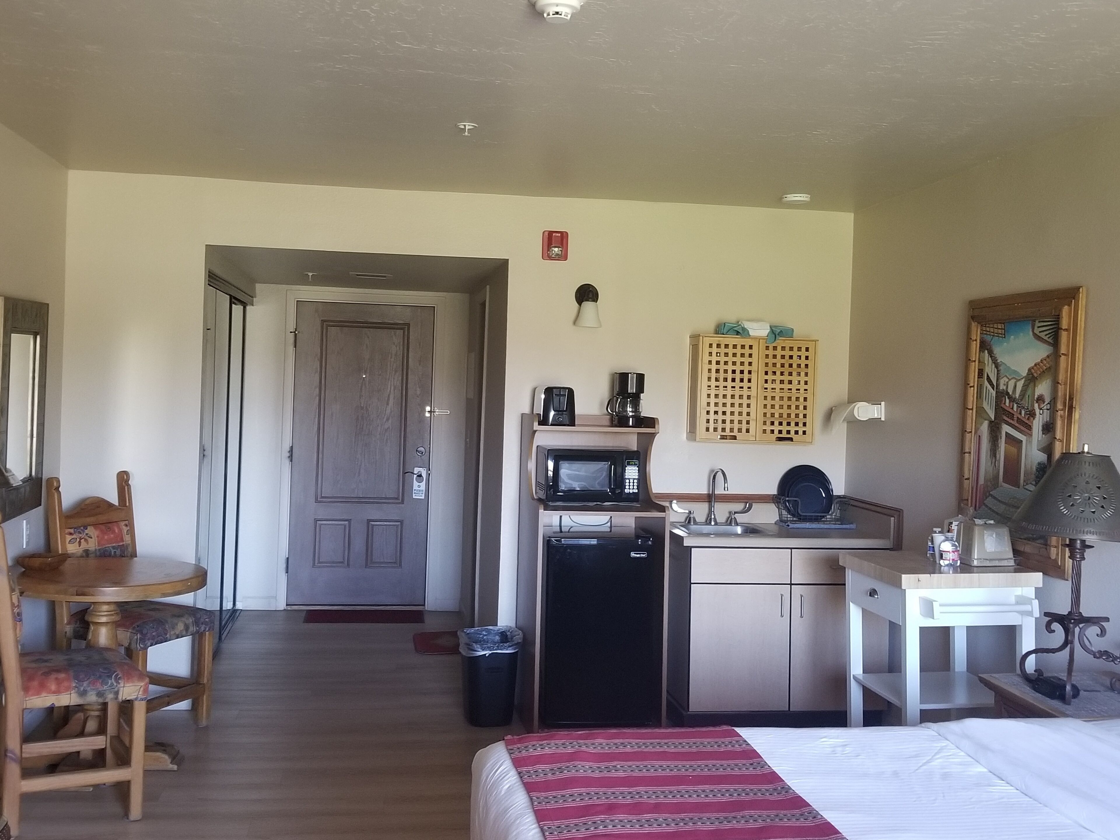 Habitació d'hotel amb un llit, una escriptori amb una cadira, una cuineta amb microones i cafetera, un rentatges i un quadre a la paret.