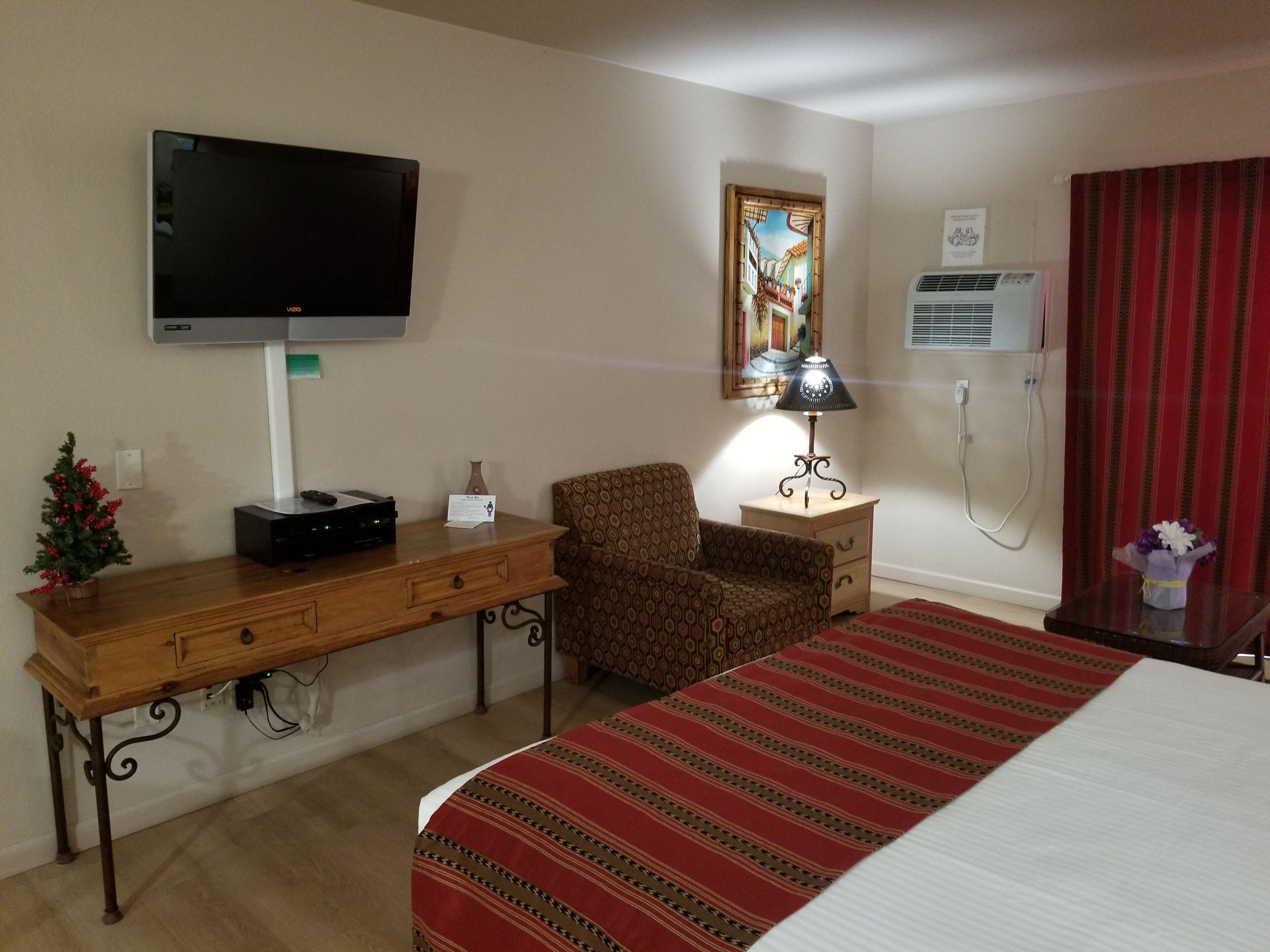 A hotel room with a bed, a desk with a flat-screen TV, a chair, a lamp, a small Christmas tree, and a painting on the wall.