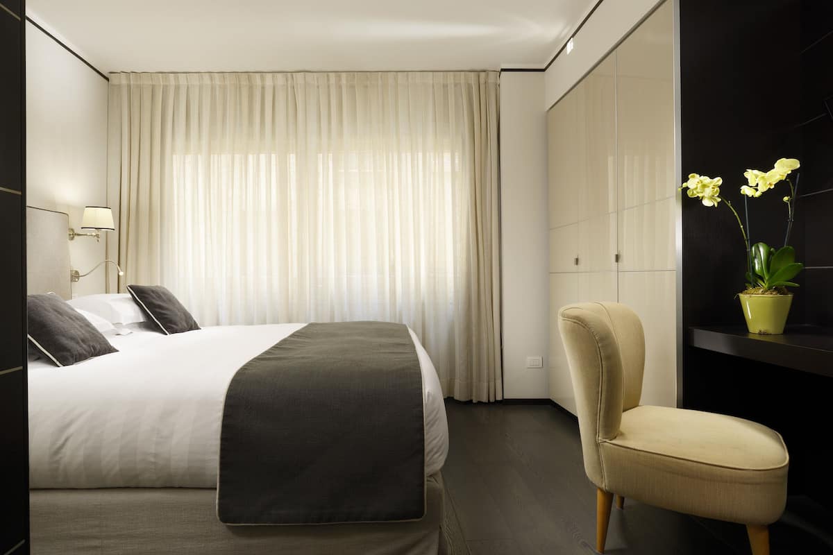 A modern hotel room with a large bed, a chair, a potted plant, and a window with sheer curtains.