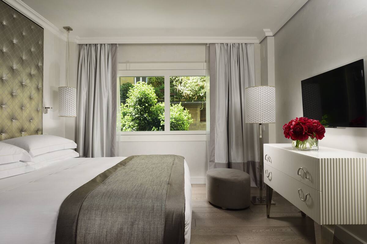 A modern hotel room with a large bed, a flat-screen TV, a bedside table with a vase of flowers, and a view of greenery outside.