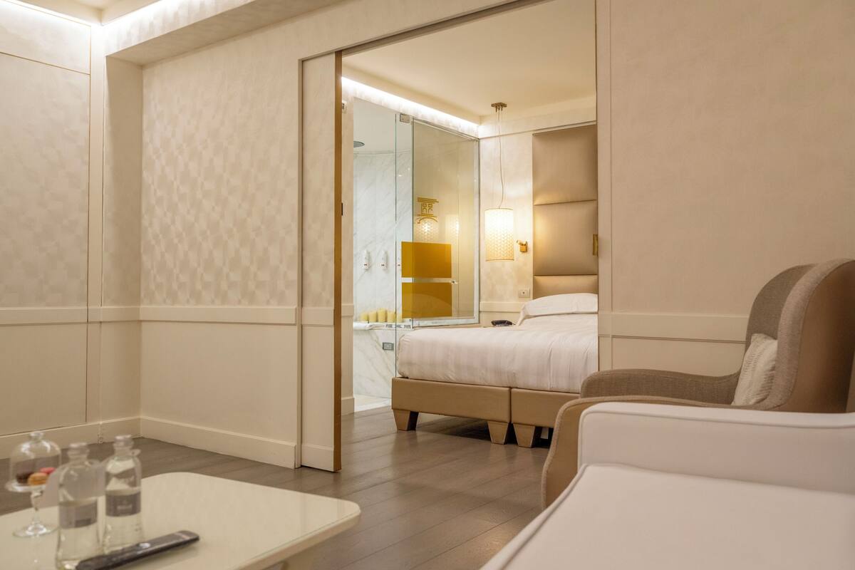 A hotel room with a bed, a sofa, a glass-enclosed shower, a bedside table with a remote control, and a mirror.