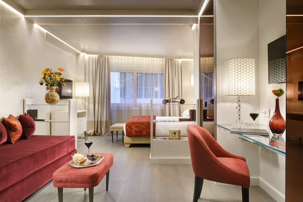 A modern hotel room with a red sofa, a glass coffee table, a red armchair, a bed with a red and white patterned bedspread, a glass desk with a lamp, and a large window with sheer curtains.