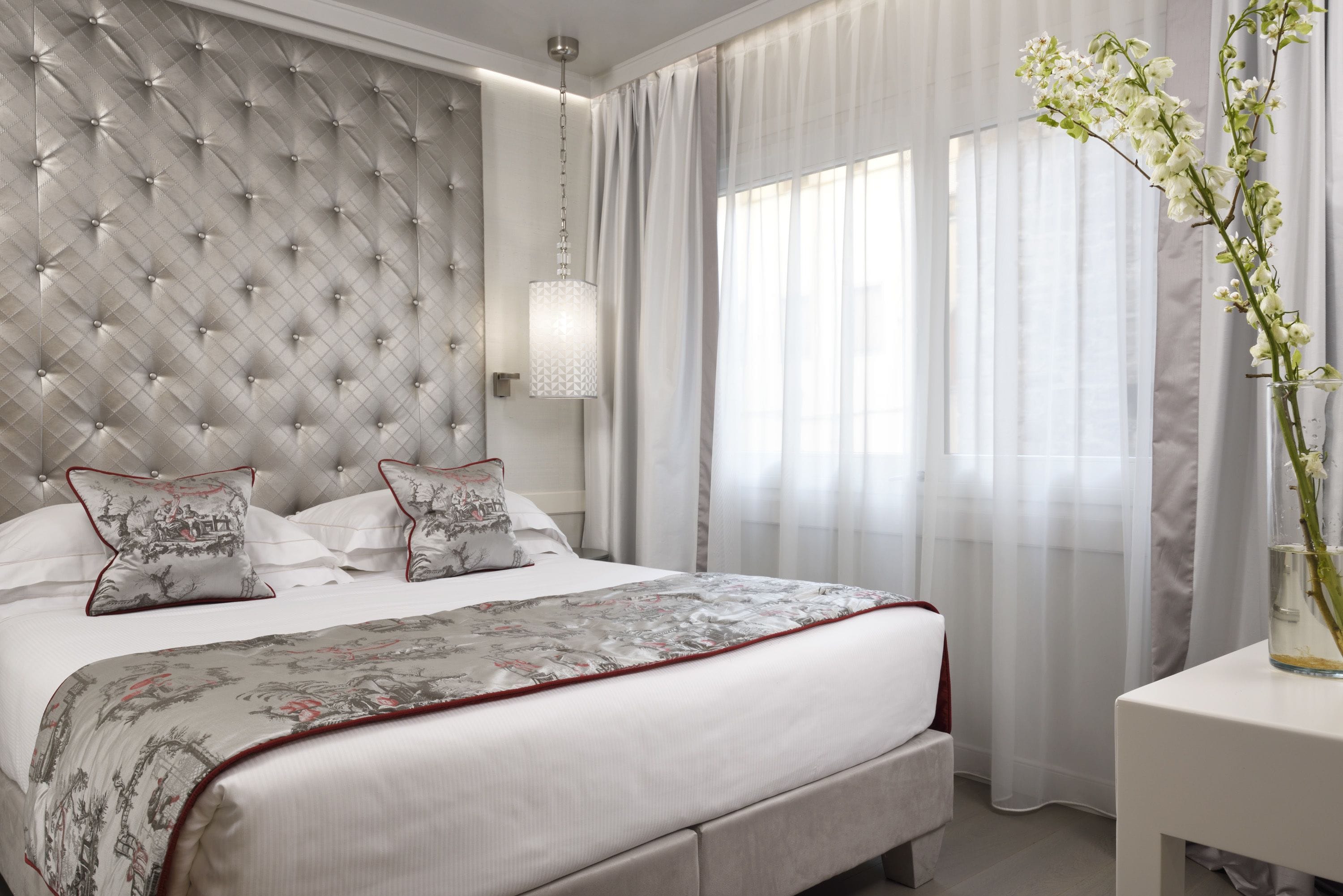 A bedroom with a tufted headboard, a bed with white linens and decorative pillows, a bedside table with a vase of flowers, and sheer curtains on the windows.