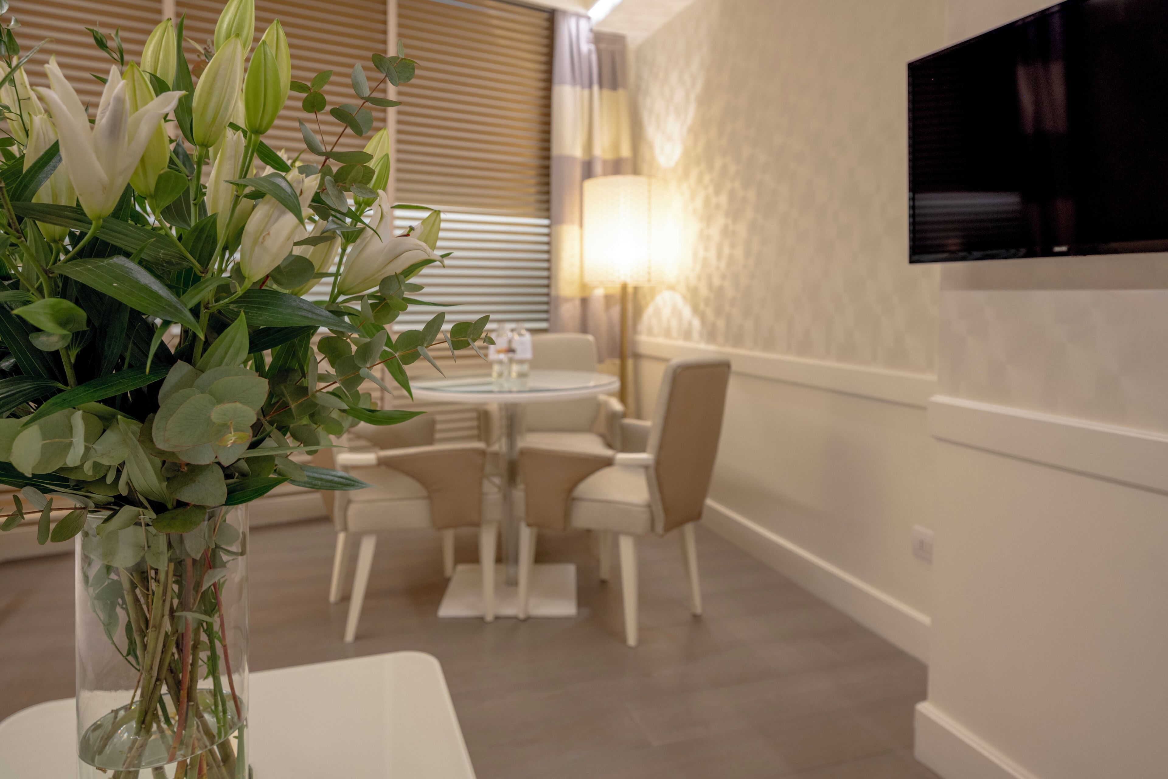 A modern dining area with a round table, chairs, a vase with flowers, a lamp, and a TV mounted on the wall.