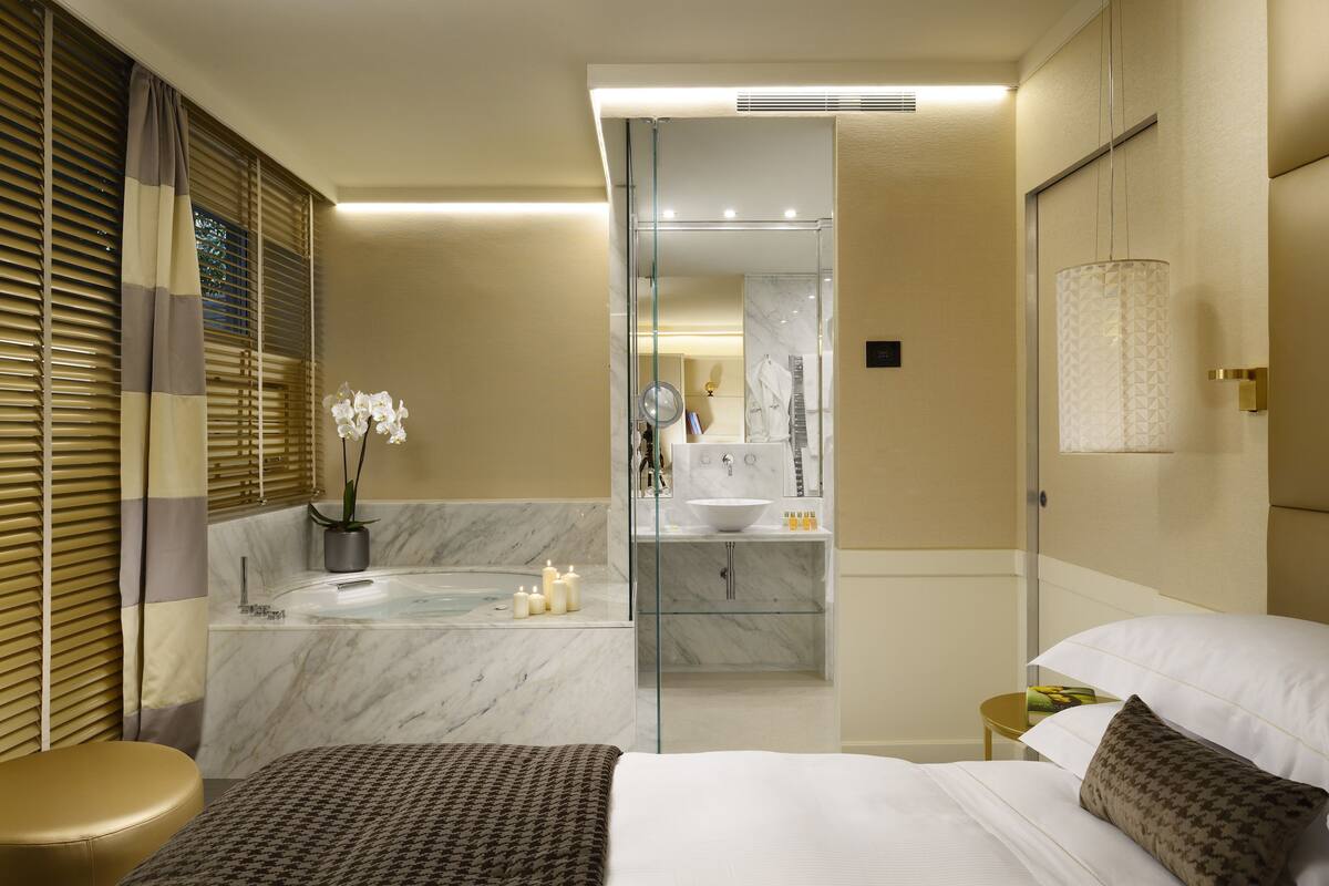 A modern hotel room with a bed, a glass-enclosed shower area, a vanity with a mirror, and a small bedside table.
