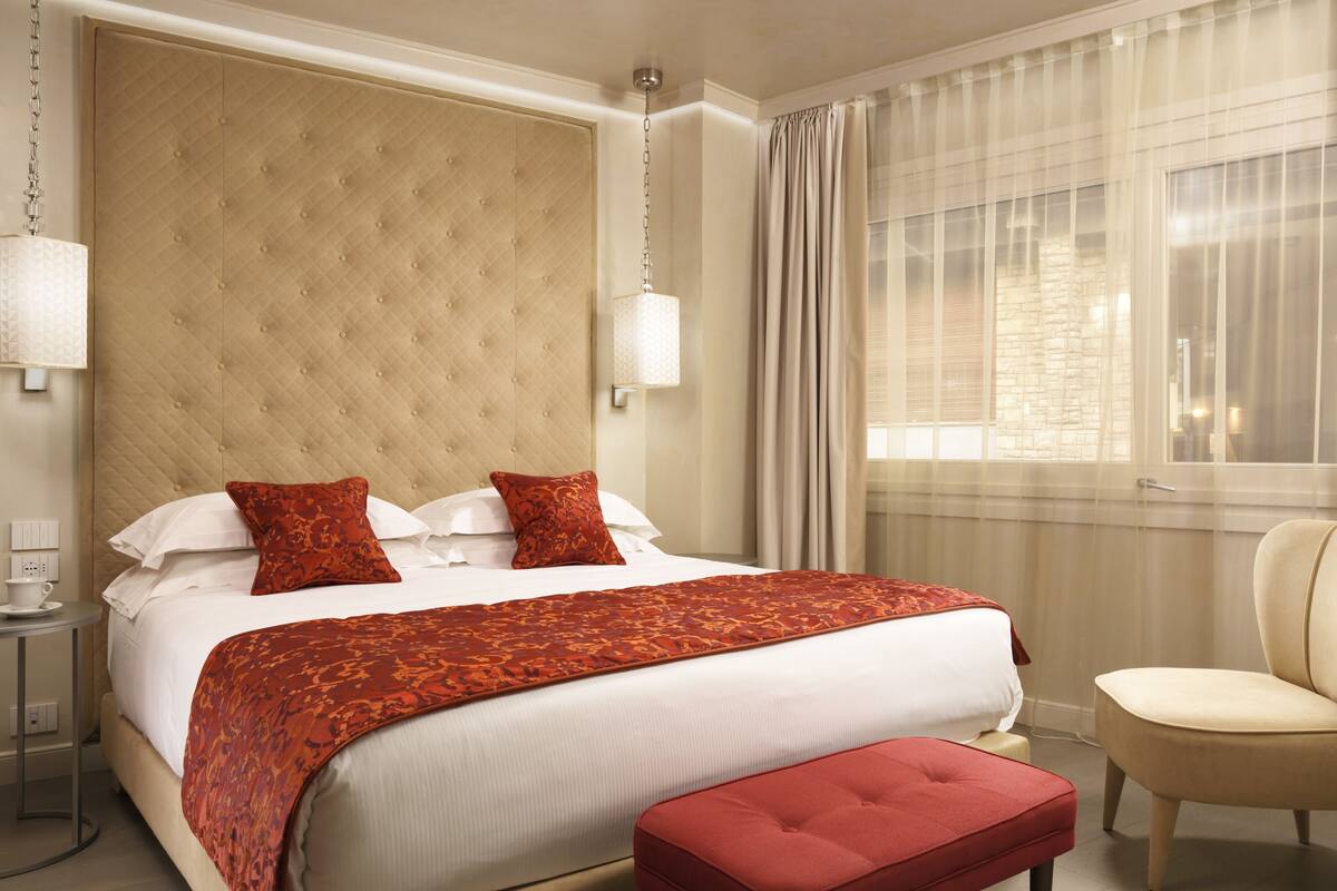 A hotel room with a large bed, a red upholstered bench, a side table, and a chair.