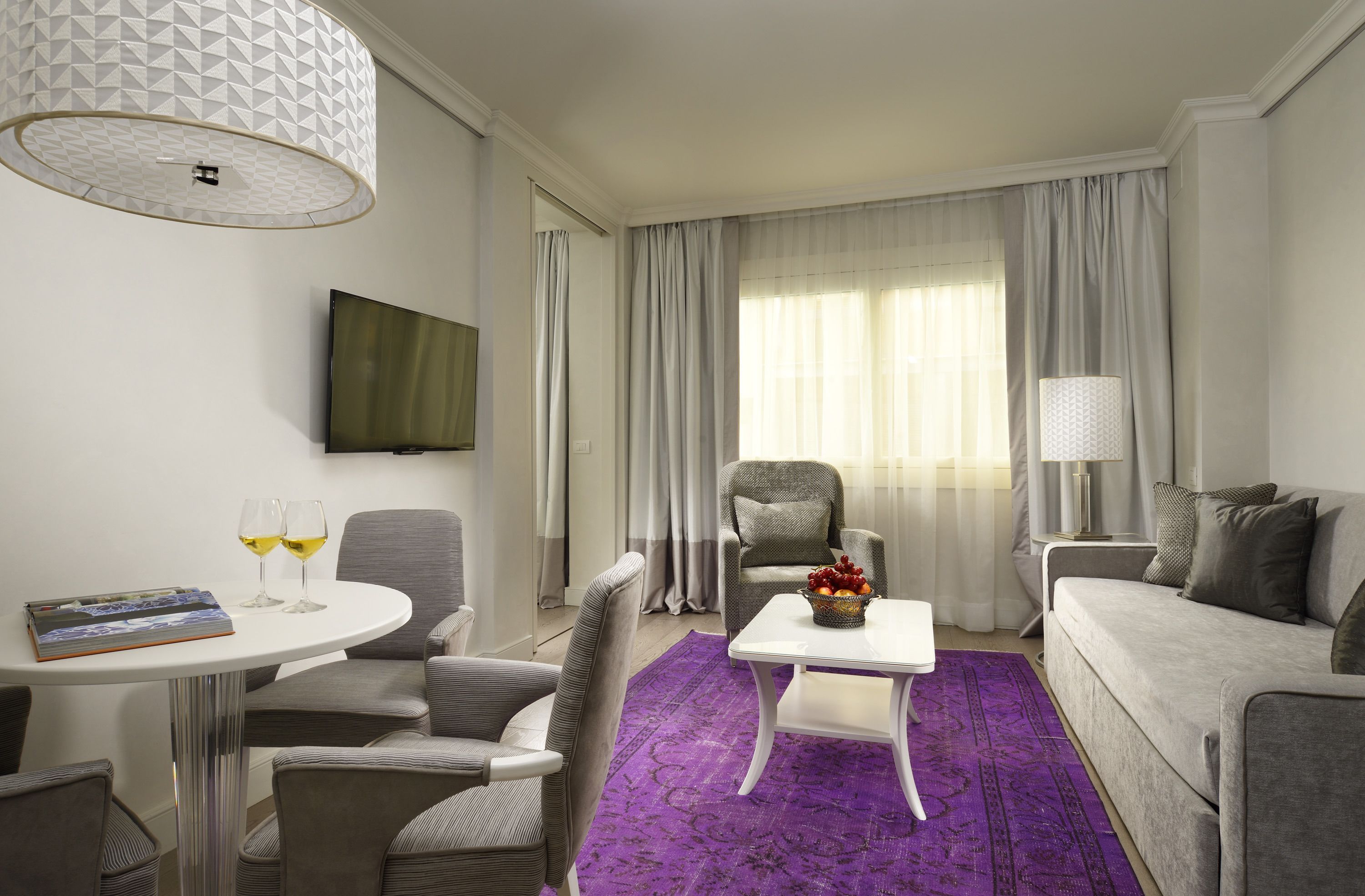 A modern hotel room with a grey sofa, a white coffee table, a purple rug, a grey armchair, a dining table with two glasses of wine, and a flat-screen TV mounted on the wall.