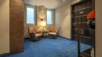 A hotel room with a blue patterned carpet, two striped armchairs, a small table, a lamp, a door with a mirror and orange roses, and two windows.