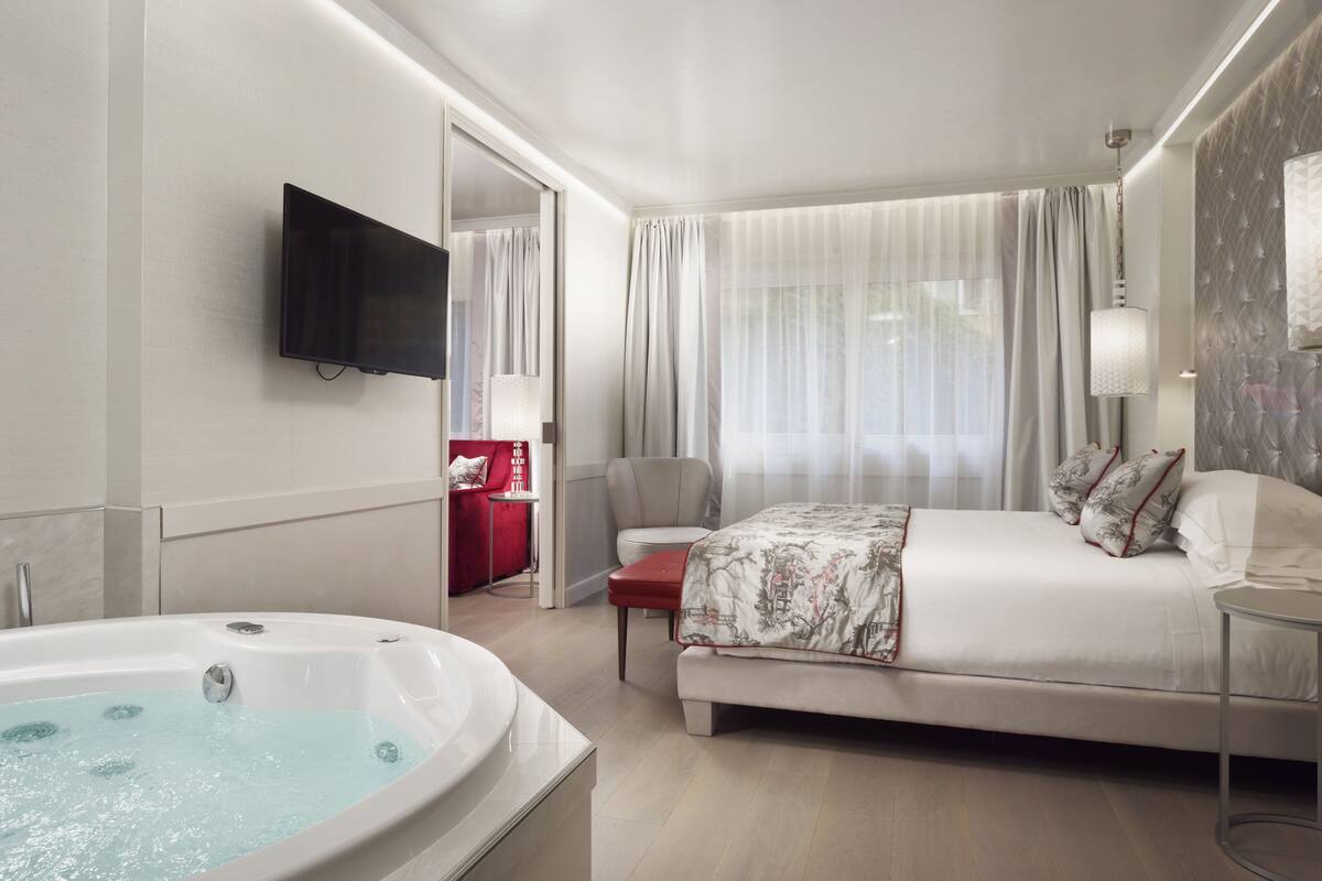 A modern hotel room with a large bed, a sitting area with a red chair, a TV mounted on the wall, and a bathtub.