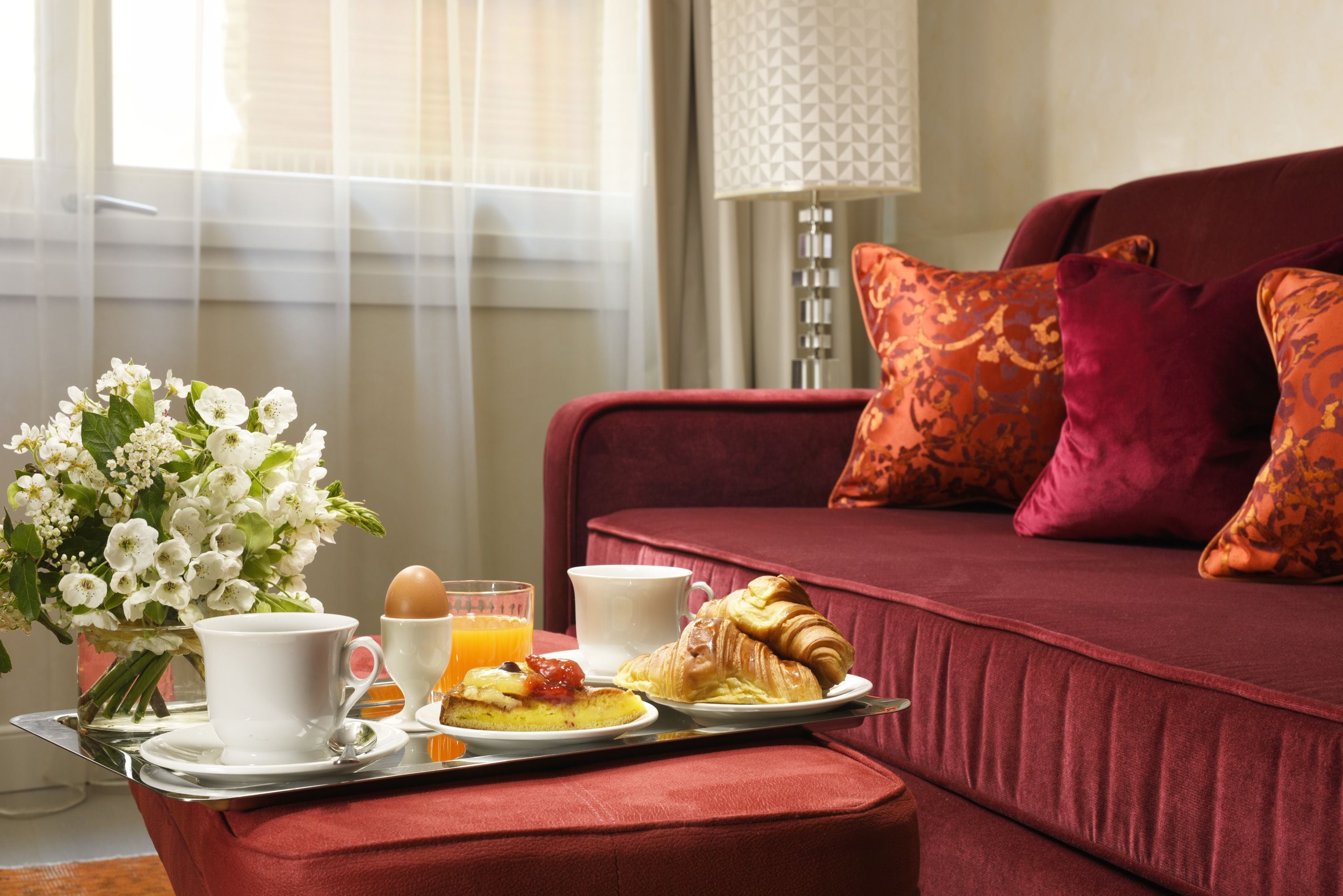 Een ontbijtkarretje met croissants, een kop koffie, een glas sinaasappelsap en een bord met eieren en toast, geplaatst op een rode gestoffeerde bank met gestreepte kussens.