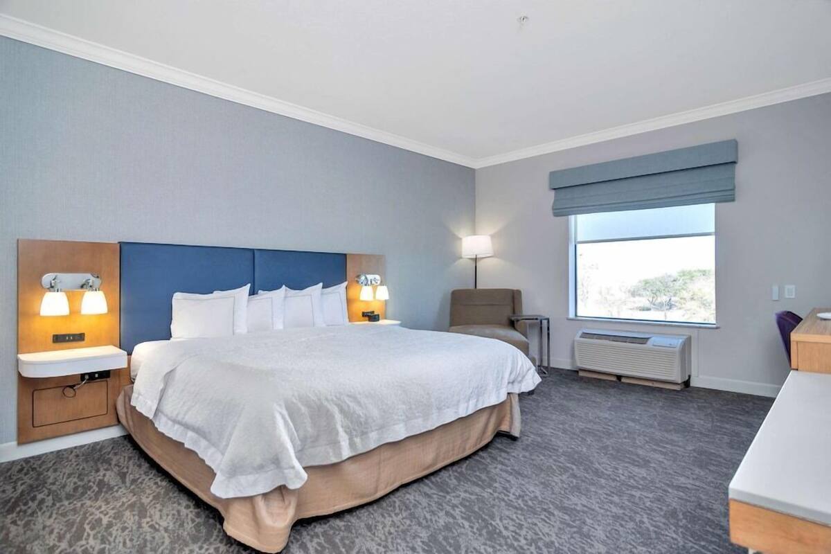 A hotel room with a large bed, bedside lamps, a window with a view, and an air conditioning unit.