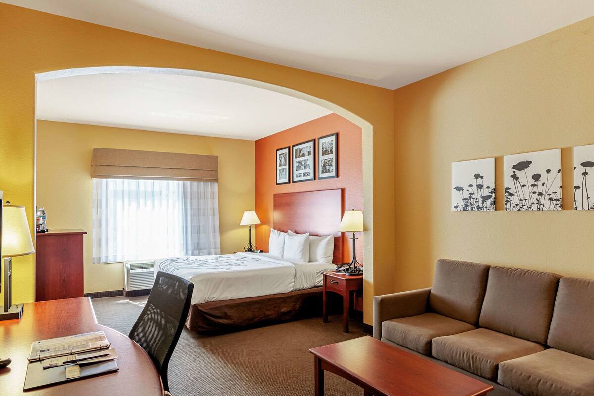 A hotel room with a bed, two bedside tables with lamps, a sofa, a desk with a chair, and artwork on the walls.