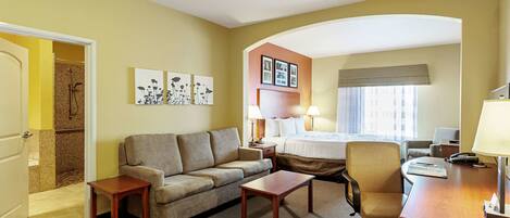Quarto de hotel com cama, sofá, escrivaninha, cadeira, mesa pequena e área de chuveiro.