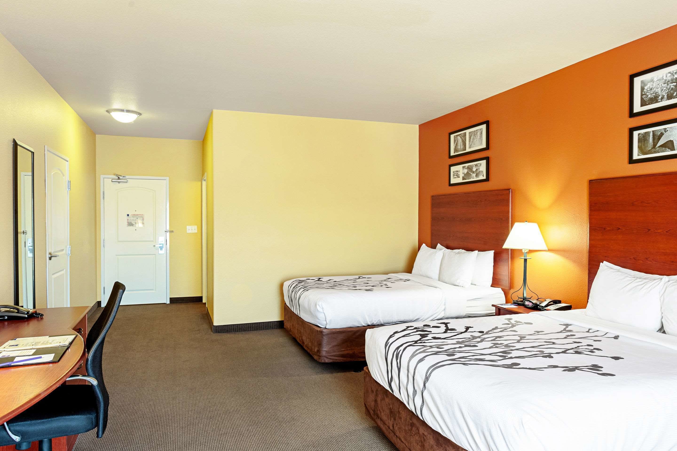 Habitación de hotel con dos camas, un escritorio y una silla. Se ve una puerta y una pared con cuadros.