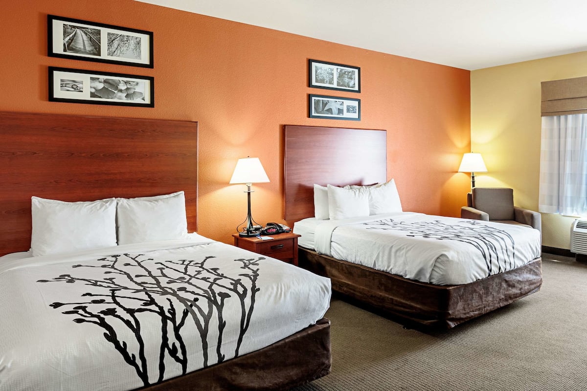 A hotel room with two beds, each with a wooden headboard, white bedding, and a lamp on a bedside table. There are framed pictures on the wall.