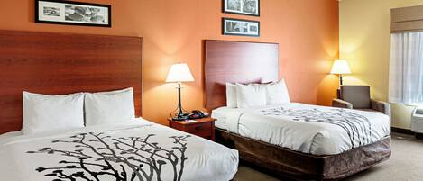 A hotel room with two beds, each with a wooden headboard, white bedding, and a lamp on a bedside table. There are framed pictures on the wall.