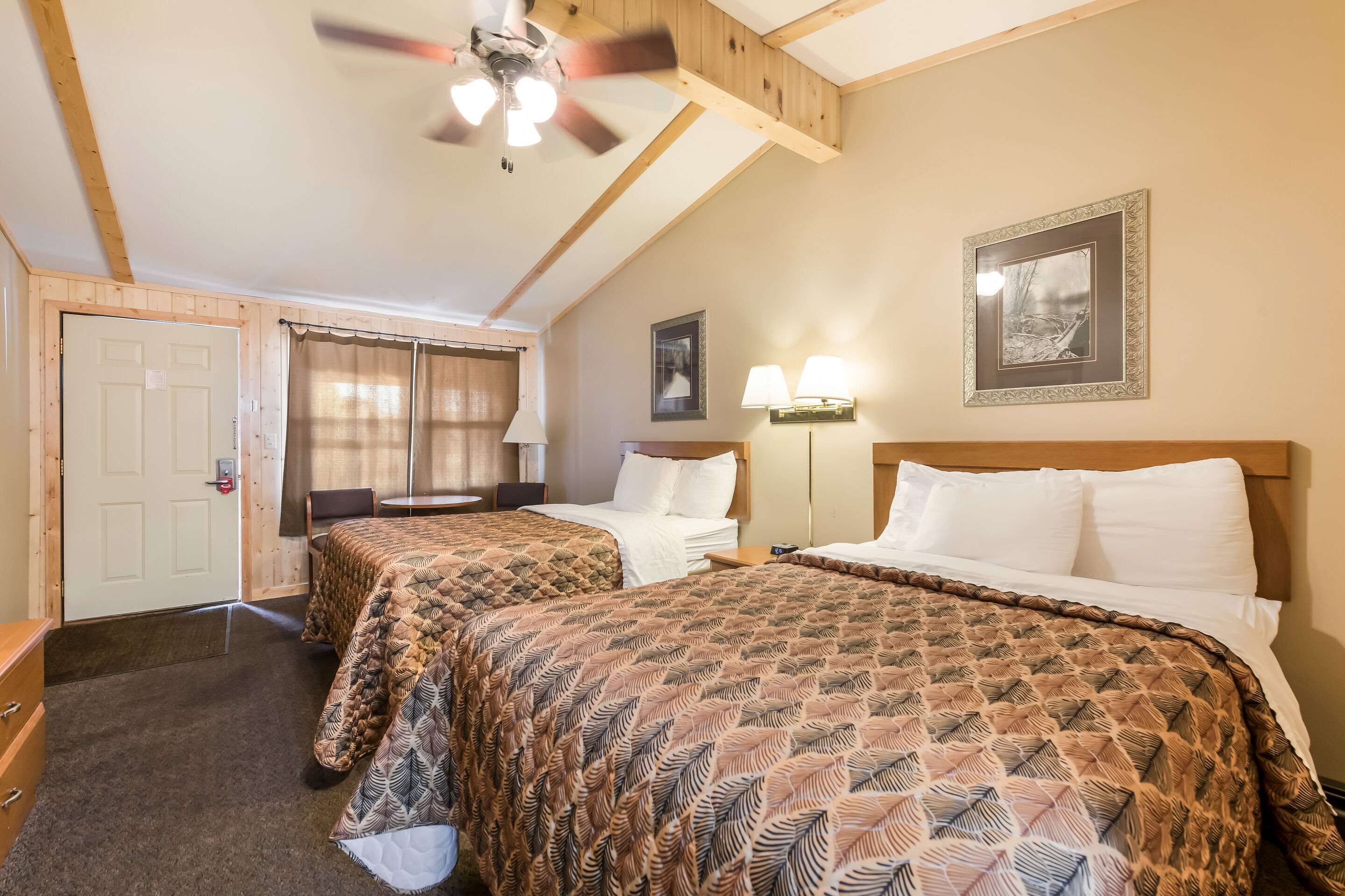 Habitación de hotel con dos camas, un ventilador de techo, una puerta, una ventana con cortinas, una mesita de noche y una lámpara fijada a la pared.