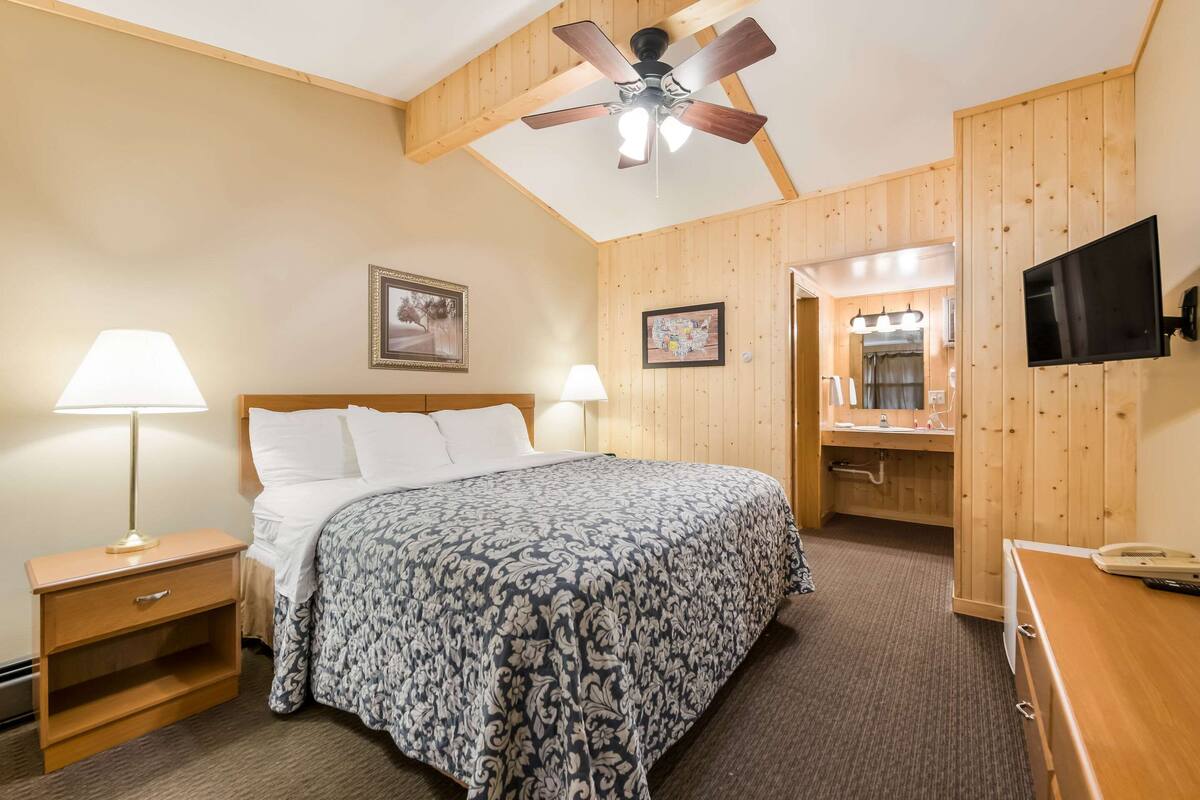 A hotel room with a bed, nightstand, lamp, ceiling fan, wooden walls, and a TV.