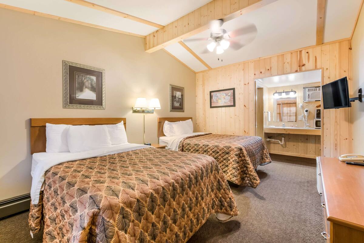 A hotel room with two beds, a ceiling fan, a TV, and a bathroom with a mirror and lights.