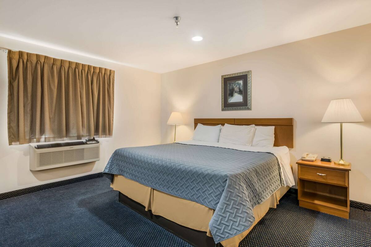 A hotel room with a bed, bedside lamps, a nightstand, a wall-mounted air conditioner, and a window with curtains.