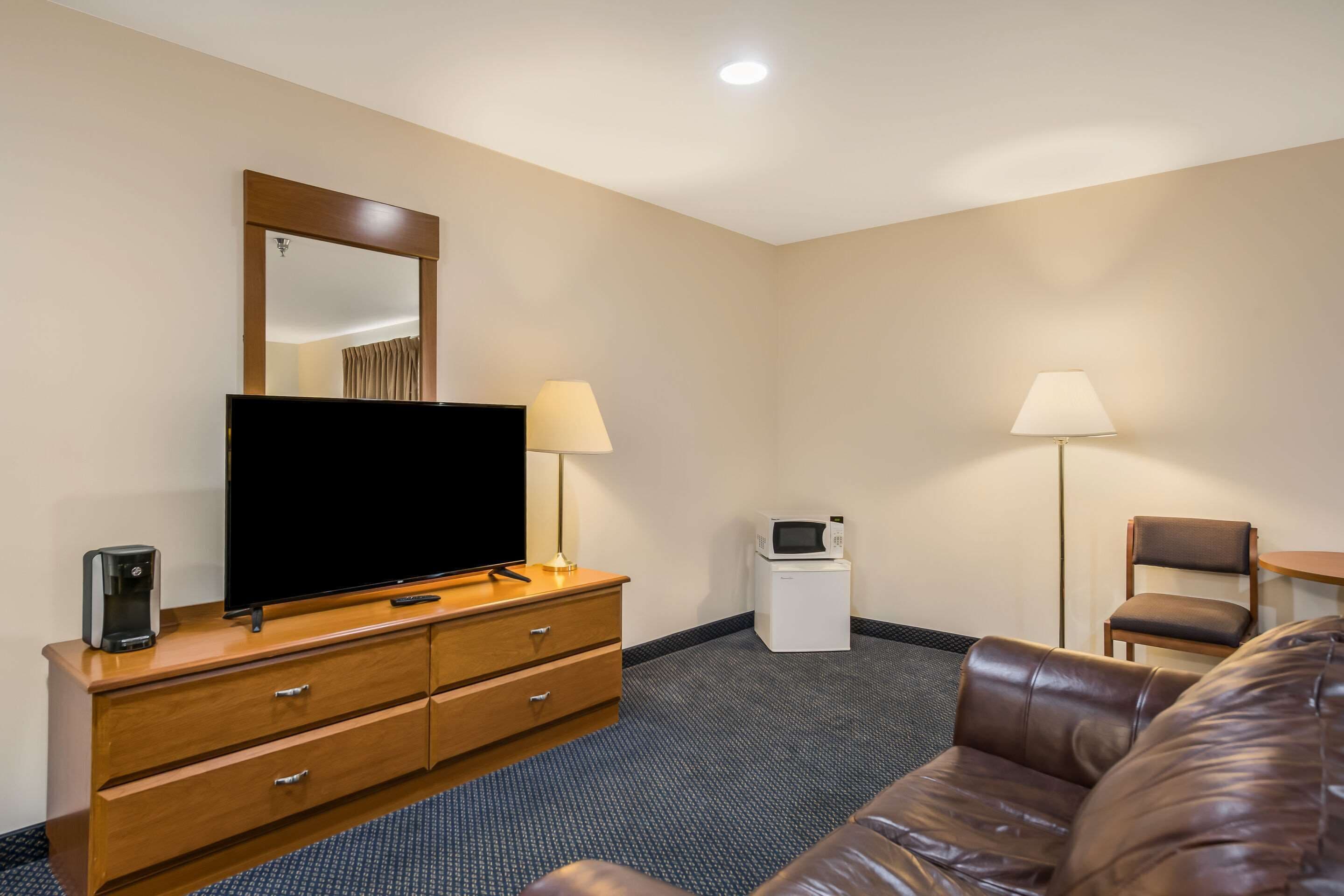 Une chambre d’hôtel équipée d’un téléviseur à écran plat, d’une commode en bois, de deux lampes, d’un four à micro-ondes, d’un canapé en cuir marron et d’une petite table.