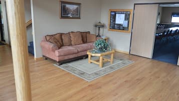 A living room with a wooden floor, a pink sofa, a small wooden table, and a lamp. There is a framed picture on the wall and a bulletin board with a welcome sign.