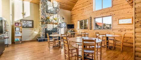 Interior espacioso de una cabaña de madera con varias mesas y sillas de comedor, una chimenea de piedra y ventanas amplias.