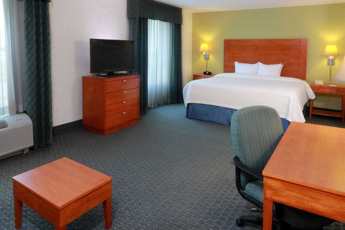 Ein Hotelzimmer mit Bett, Schreibtisch, Stuhl, Fernseher auf einer Kommode und Lampe auf dem Nachttisch.