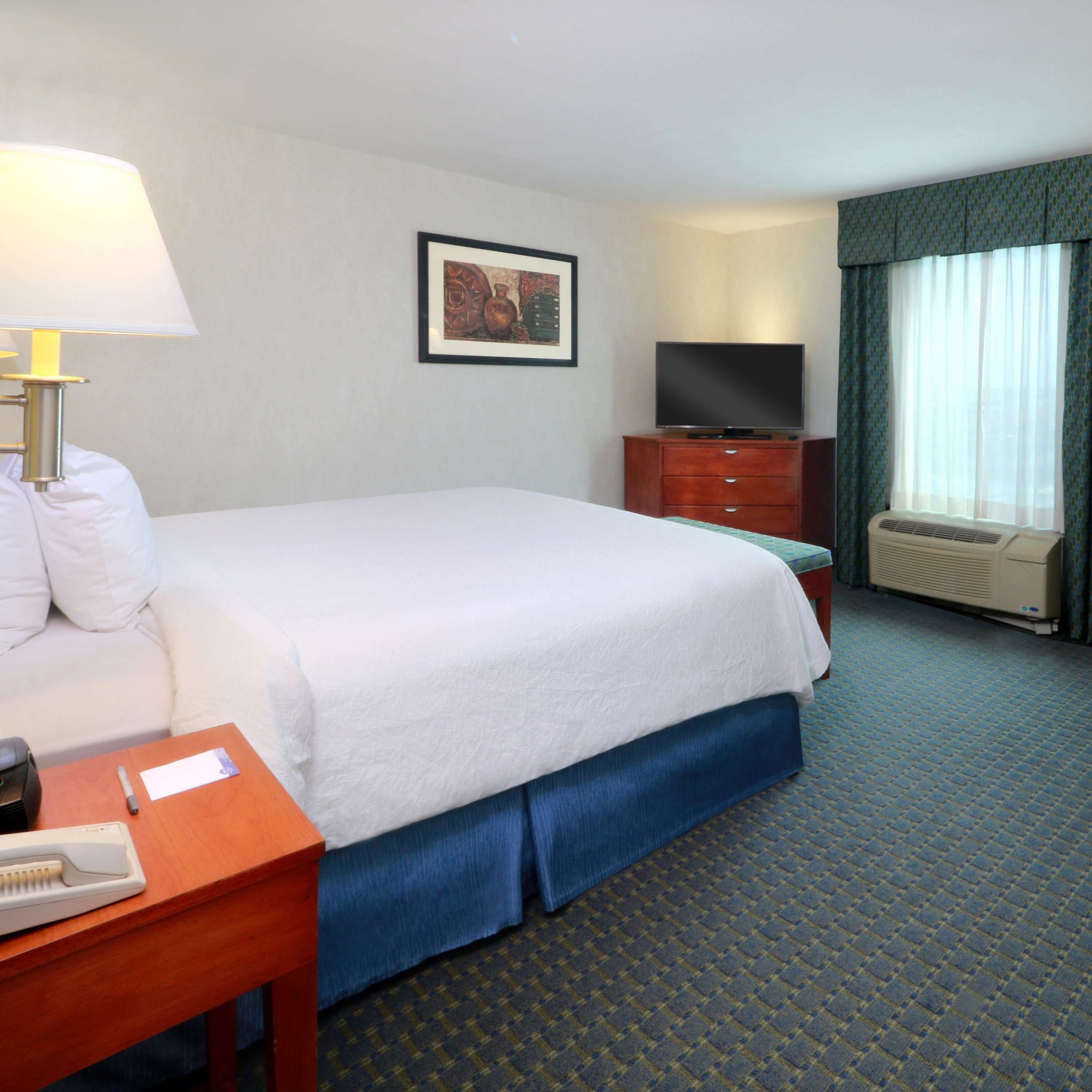 Habitación de hotel con cama, escritorio con lámpara, televisión y ventana con cortinas.
