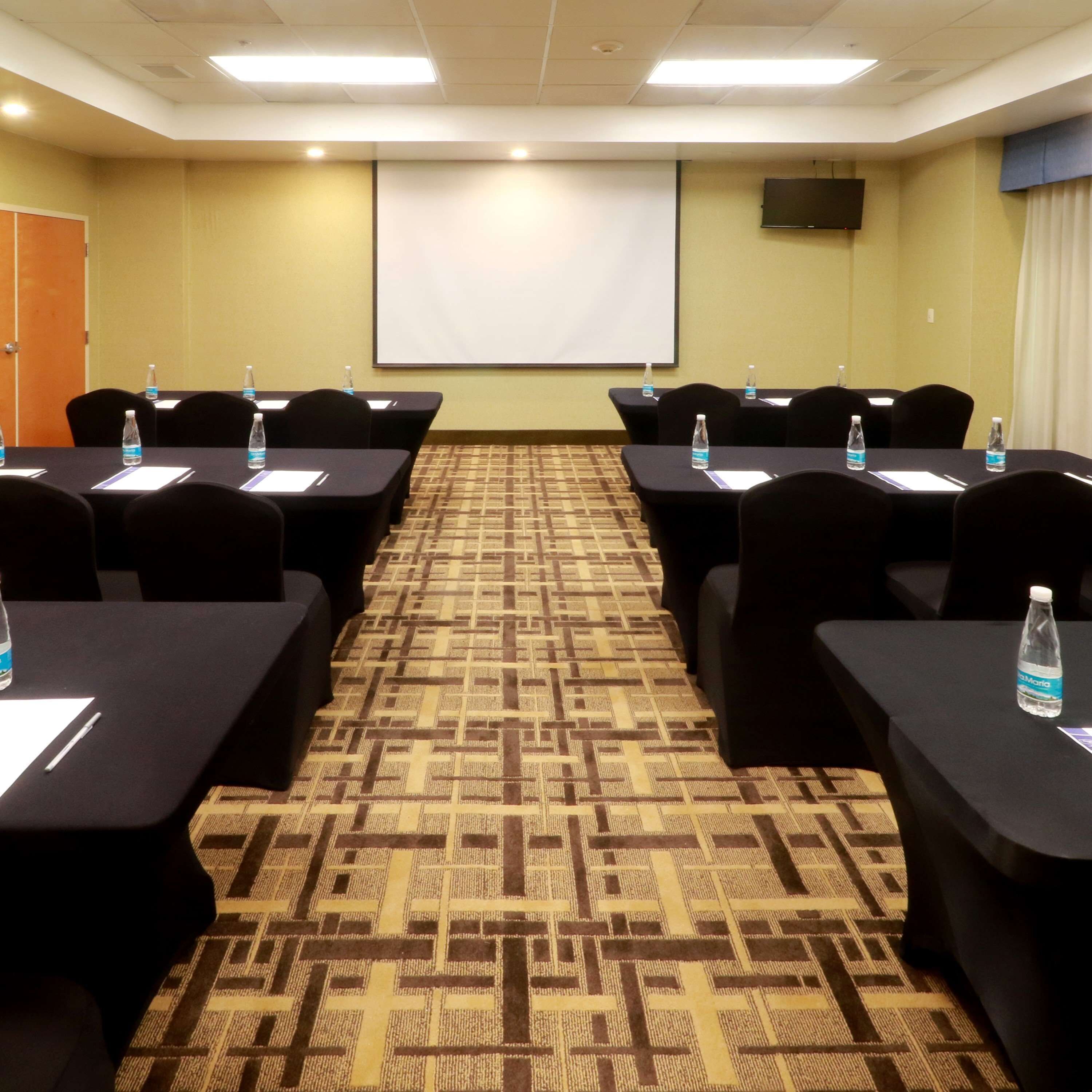 Un salón de conferencias con alfombra estampada, mesas negras con botellas de agua y una pantalla de proyector en la pared.
