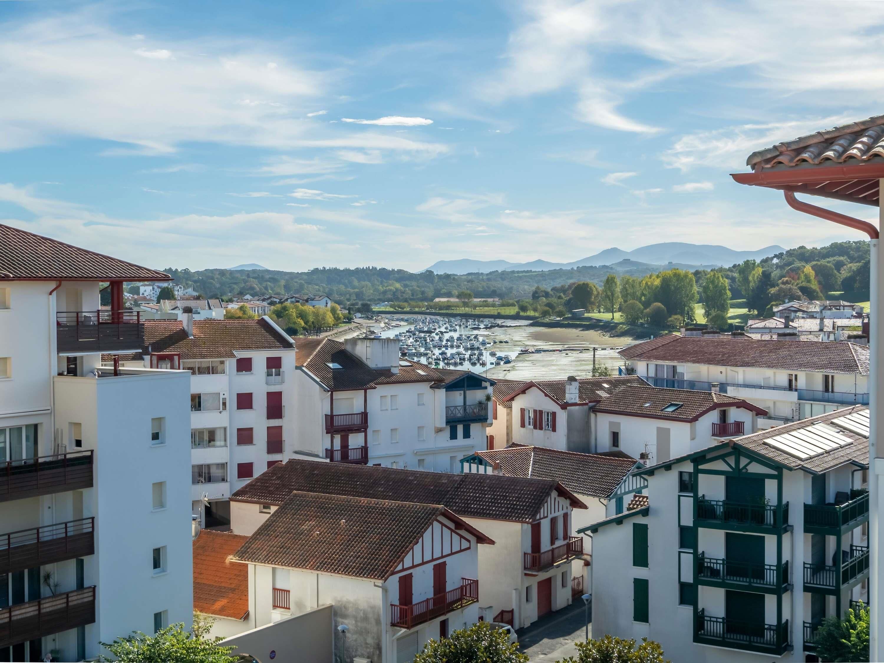 A coastal town with white buildings, red-tiled roofs, and a marina in the background.
