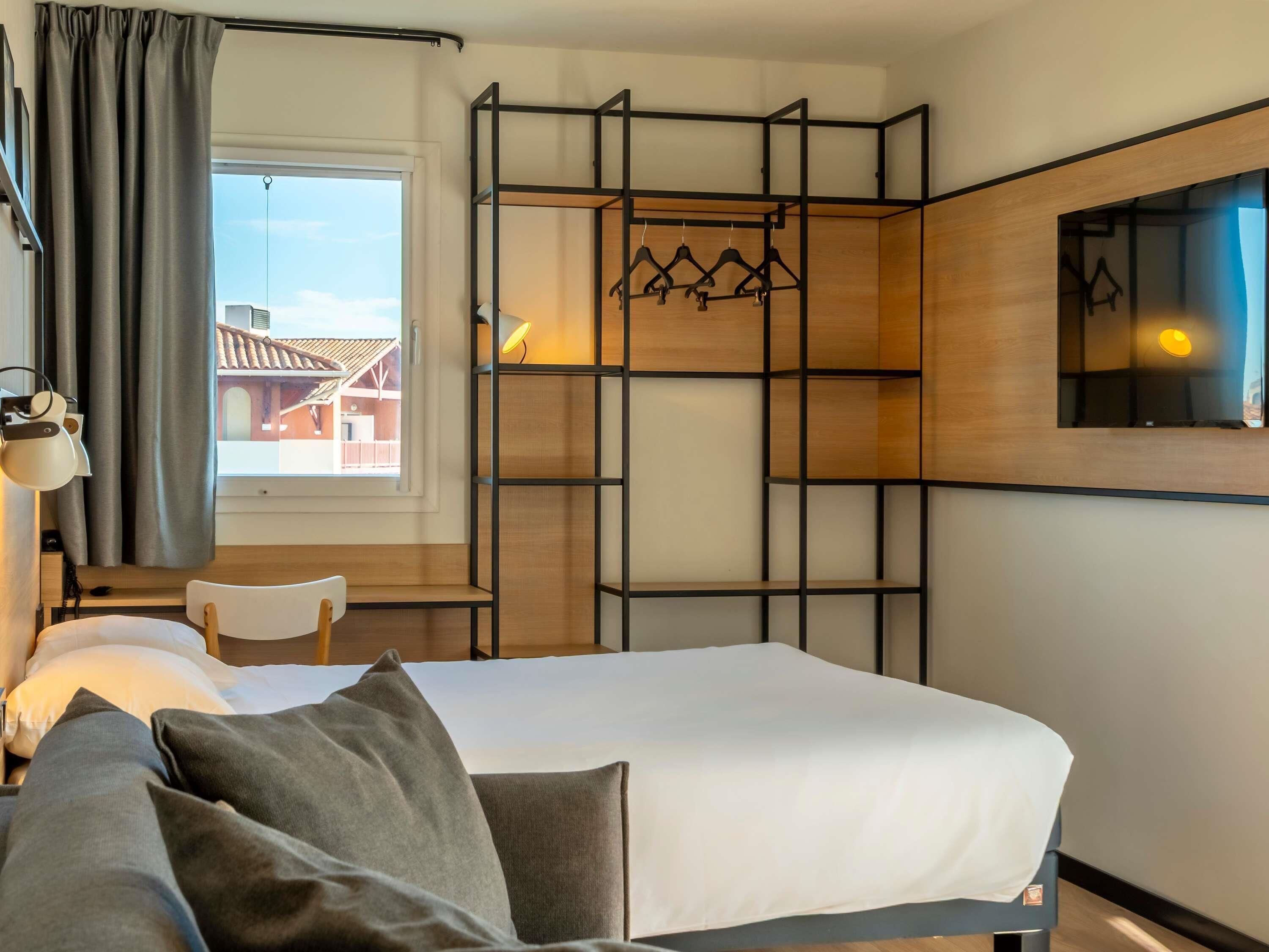 A hotel room with a bed, a desk, a chair, a wardrobe with hanging space, and a window with curtains.
