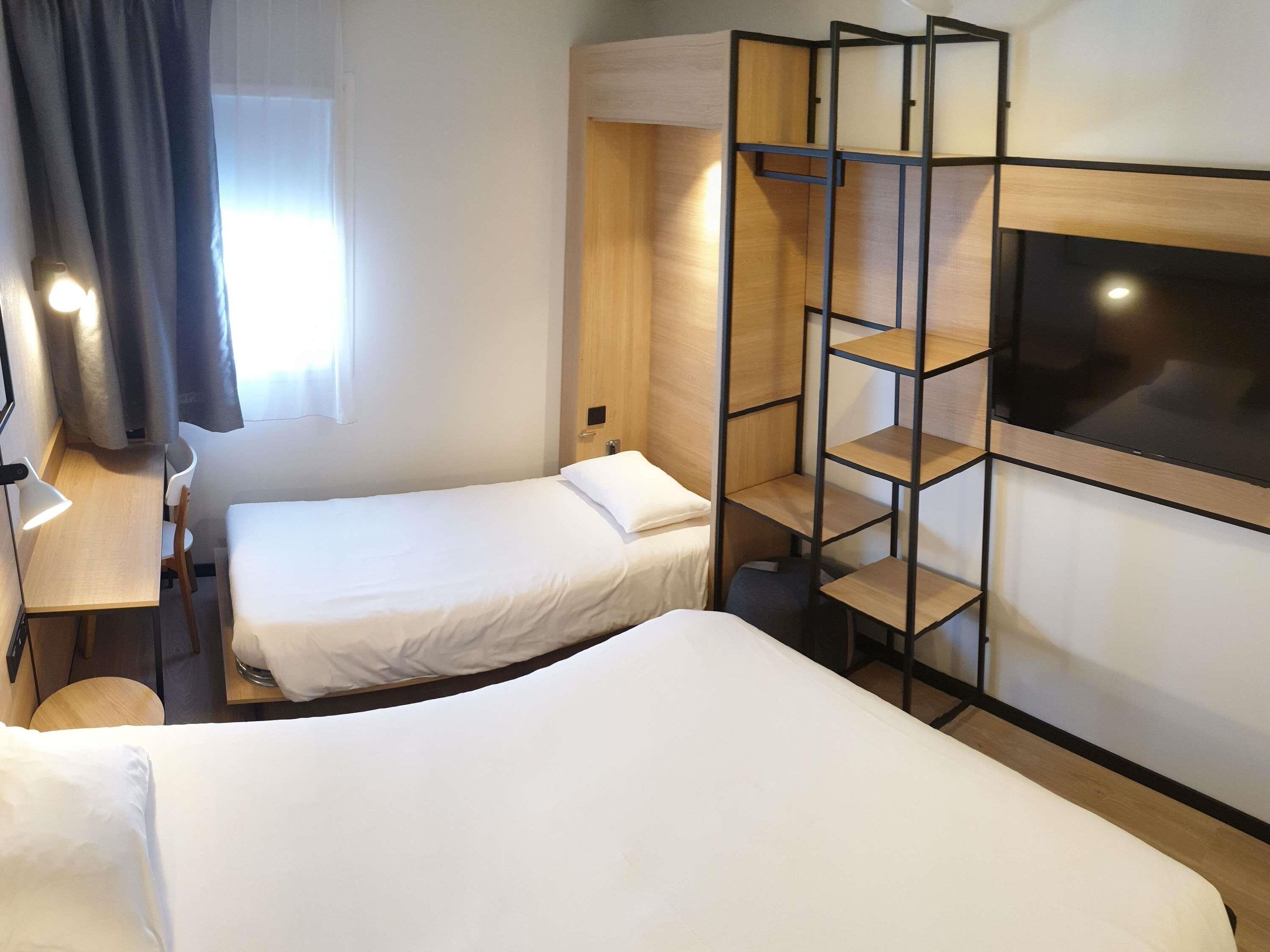 A hotel room with two single beds, a desk with a chair, a TV mounted on the wall, and a window with curtains.