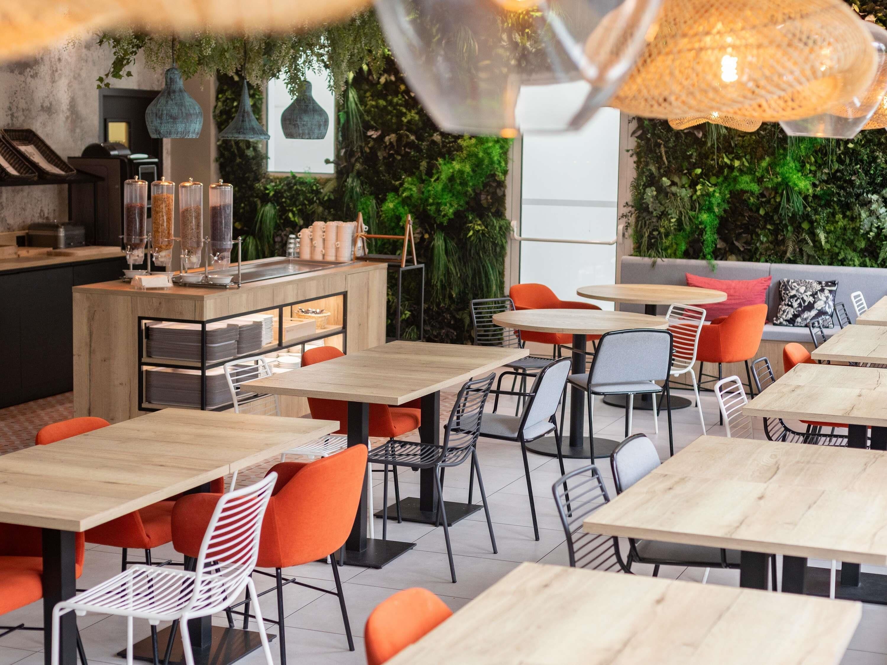 Un café al aire libre con mesas de madera y sillas color naranja, rodeado de follaje y una zona de cocina moderna.