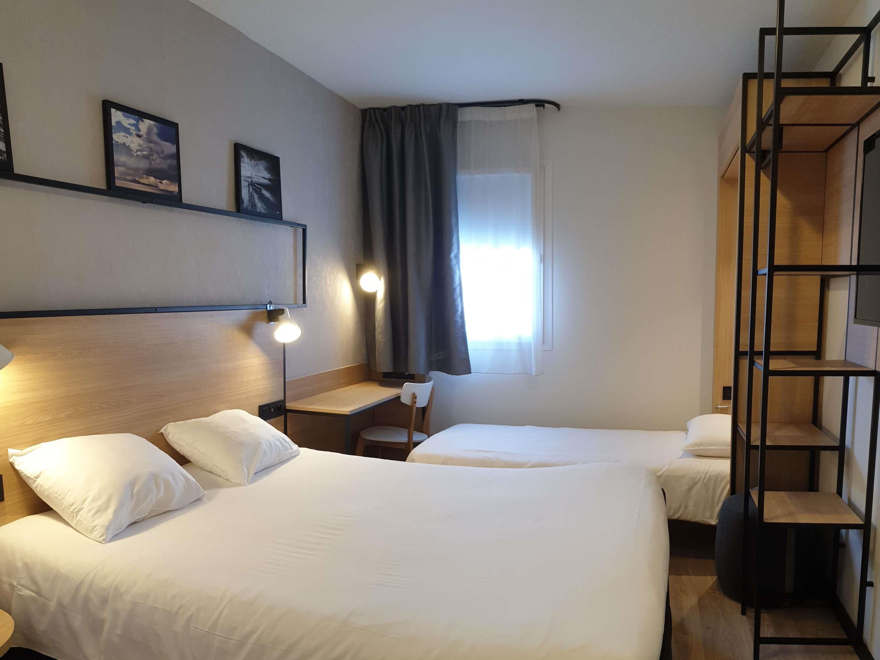 Una habitación de hotel con dos camas, un escritorio, una silla, una lámpara y un ventanal con cortinas.