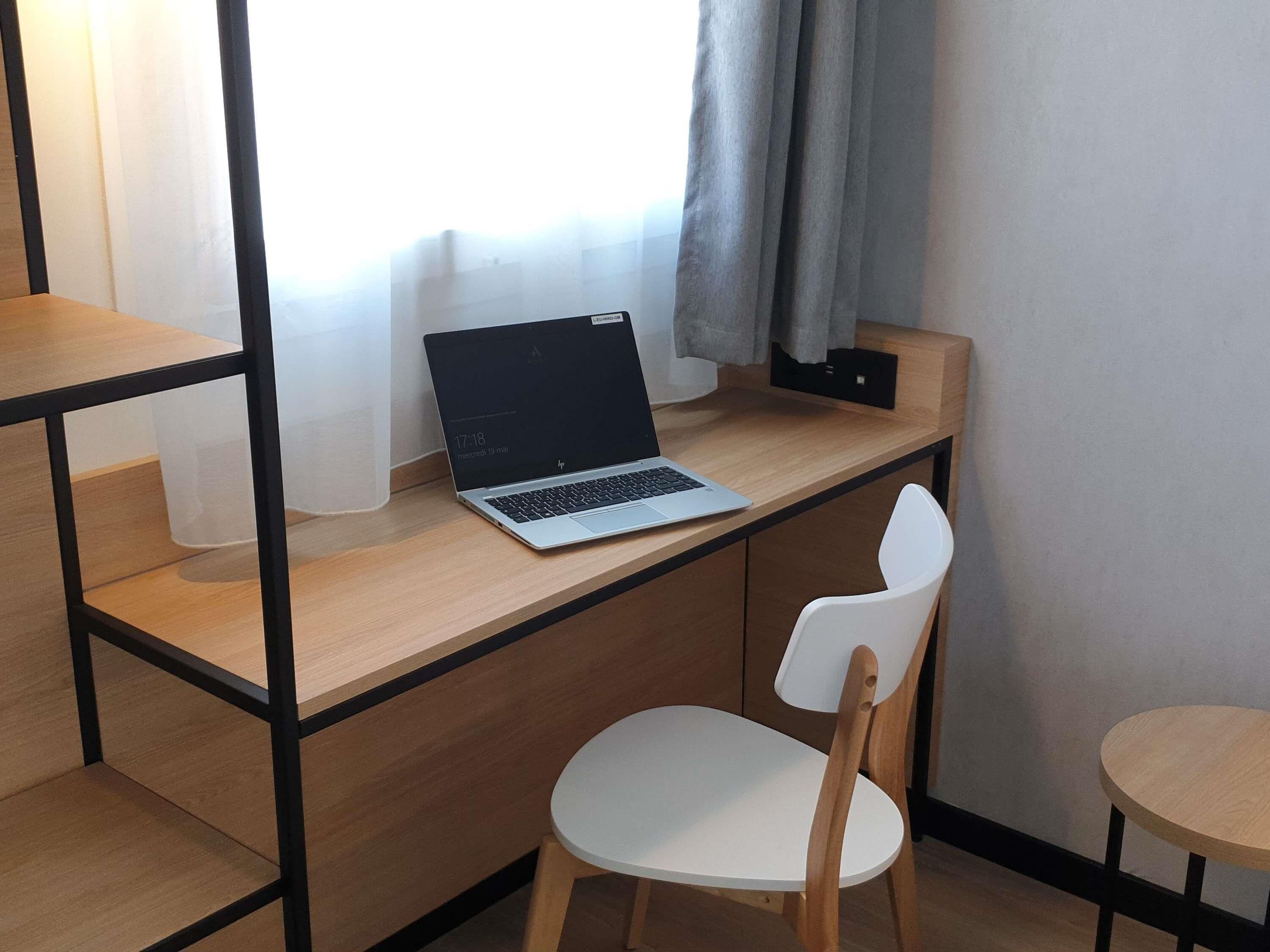 Een laptop op een houten bureau met een witte stoel, een klein rond tafeltje en een raam met gordijnen.
