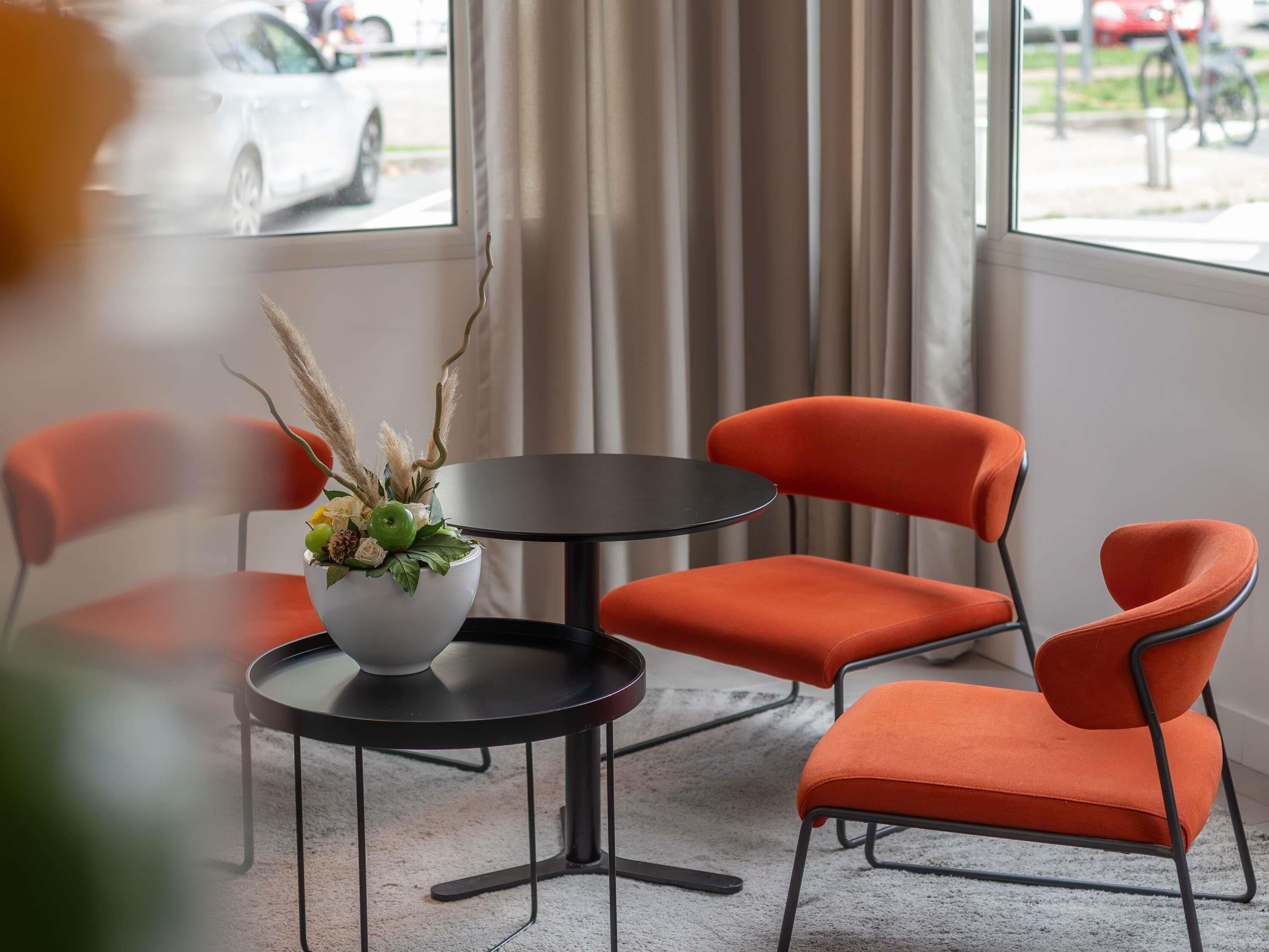 A modern room with a round table and two orange chairs, a potted plant on the table, and a view of the street through large windows.