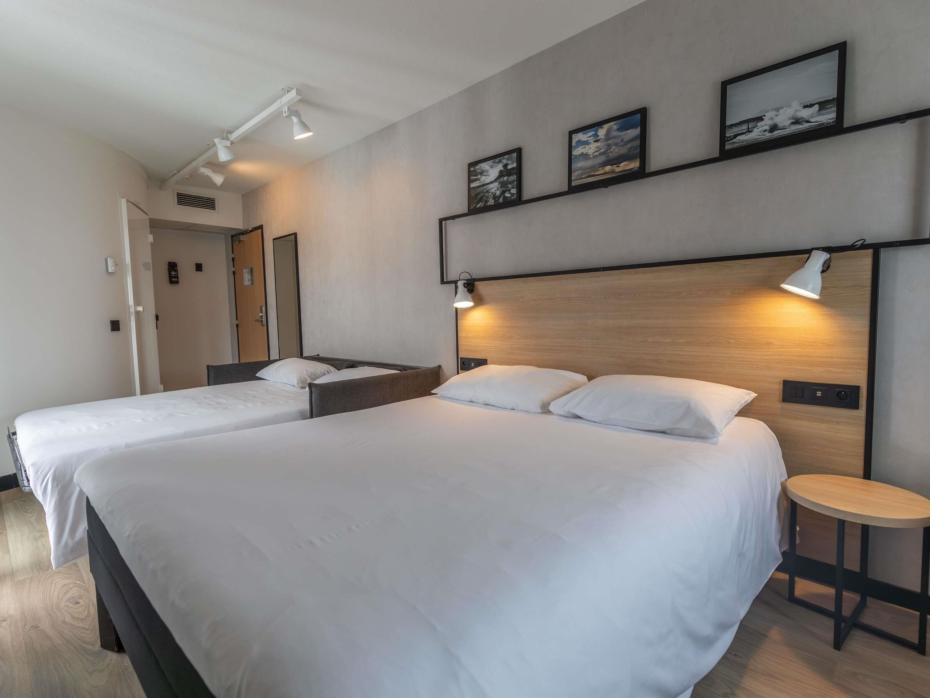 Habitación de hotel con dos camas, cabecero de madera, lámparas de noche, una mesita redonda y un estante con fotografías.