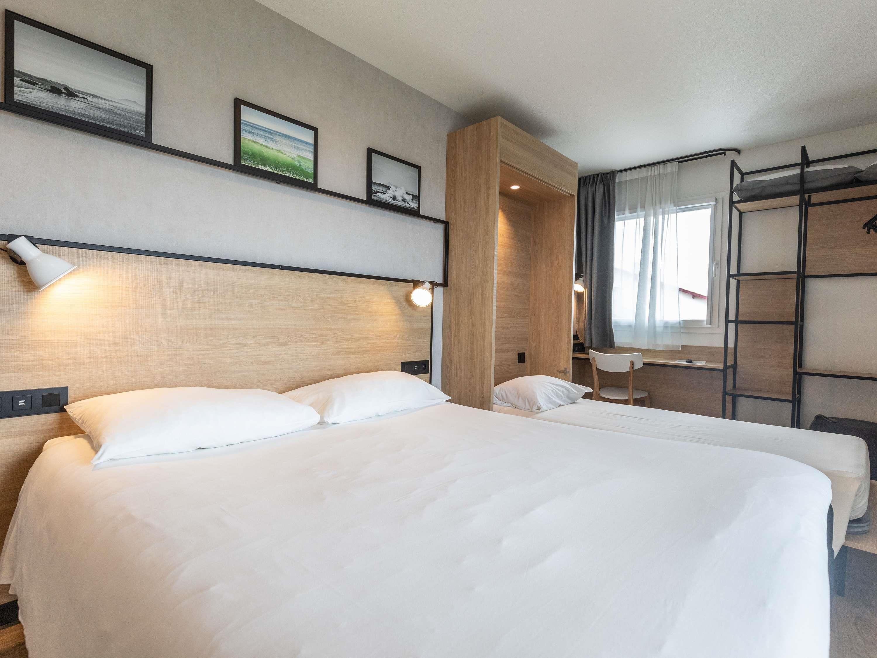 A hotel room with two beds, a wooden wardrobe, a desk with a chair, and a window with curtains.