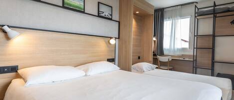 A hotel room with two beds, a wooden wardrobe, a desk with a chair, and a window with curtains.