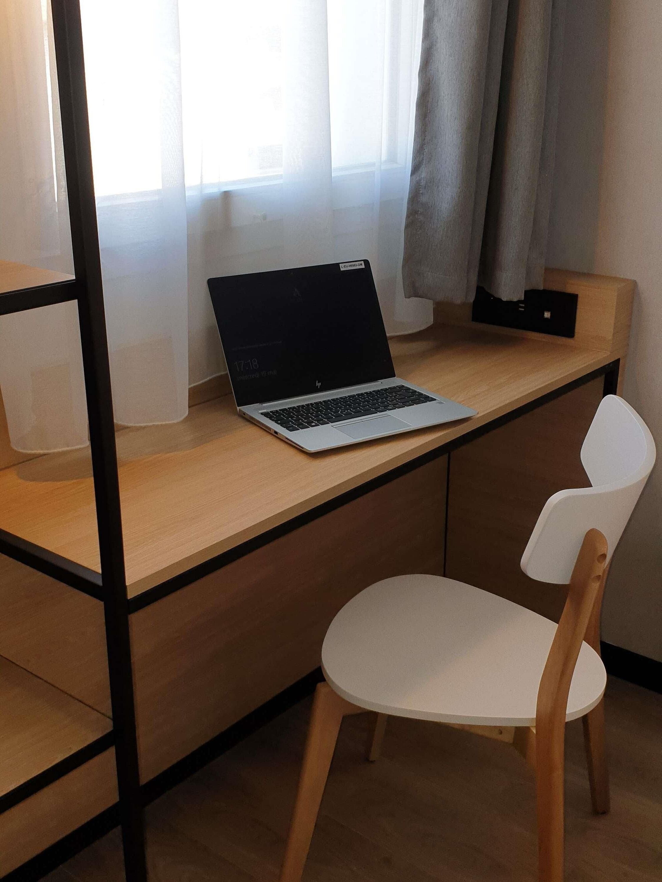 Un espacio de trabajo moderno con una laptop sobre un escritorio de madera, una silla blanca y una ventana con cortinas de gasa.