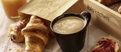 Una bandeja de madera con croissants, una taza negra con una bebida cremosa y una bolsa de papel con texto.