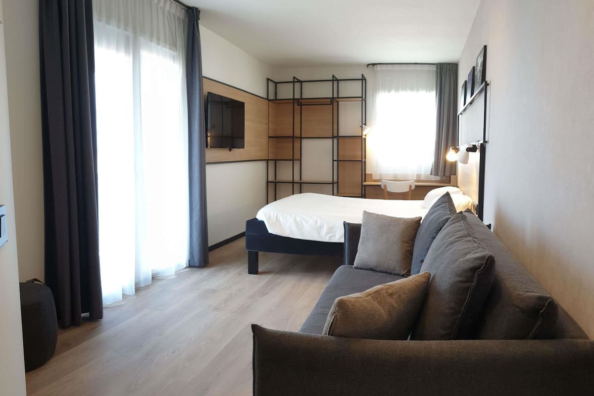 A modern hotel room with a bed, a sofa, a TV, and a desk with a lamp.