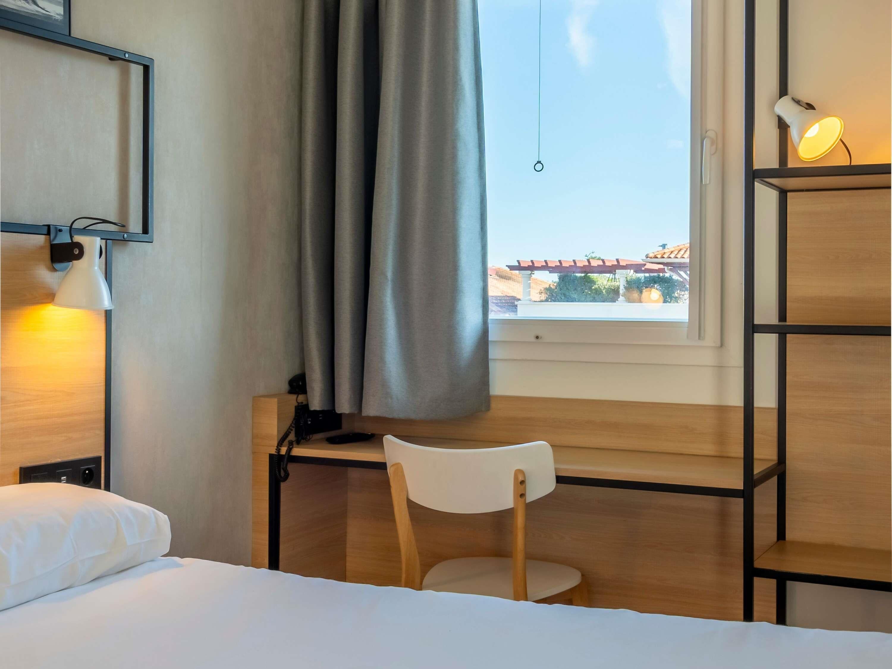 A hotel room with a bed, a desk, a chair, a bookshelf, a window with curtains, and a hanging light fixture.