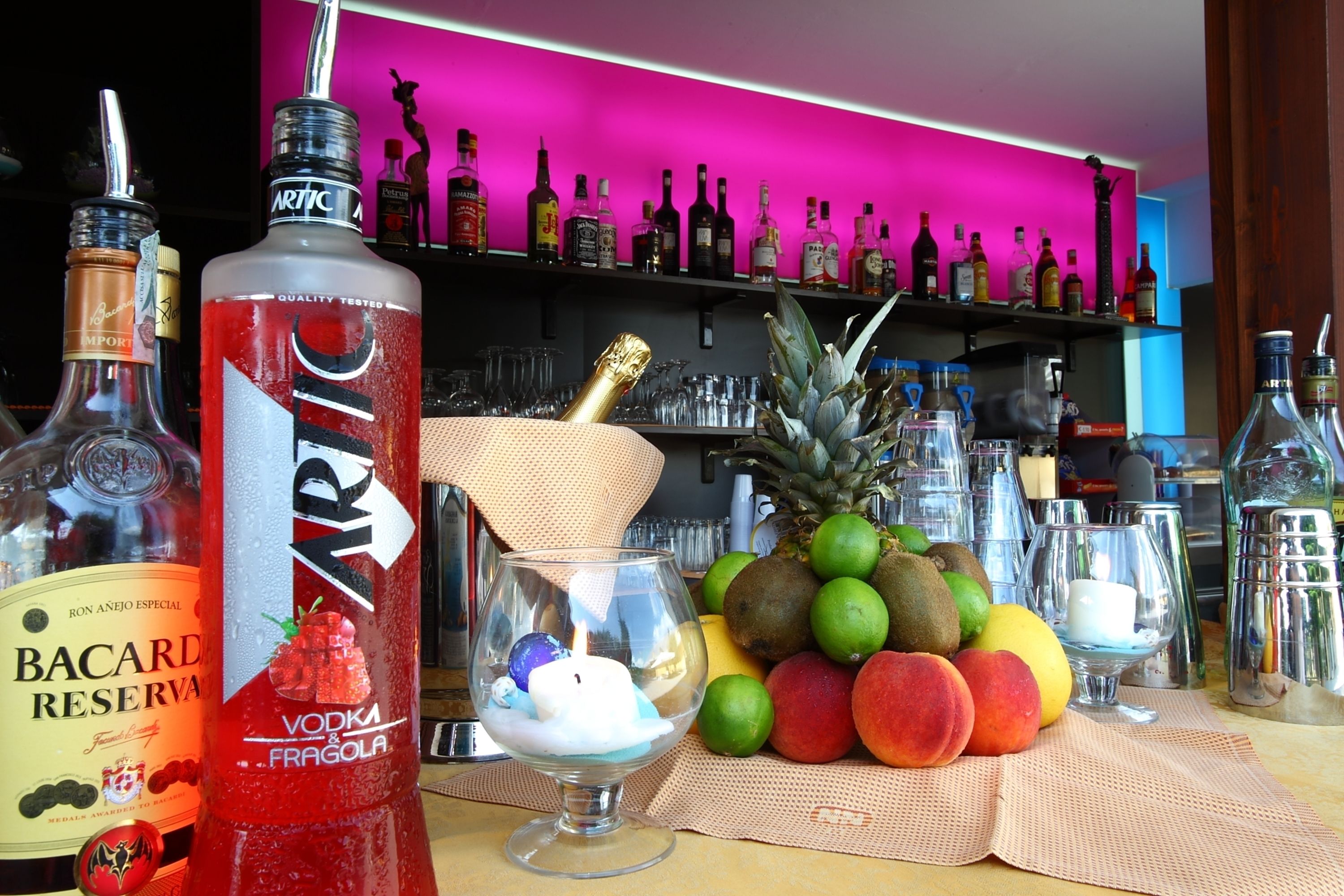 Angolo bar con diverse bottiglie di liquori, un espositore di frutta e un bicchiere con una bevanda.