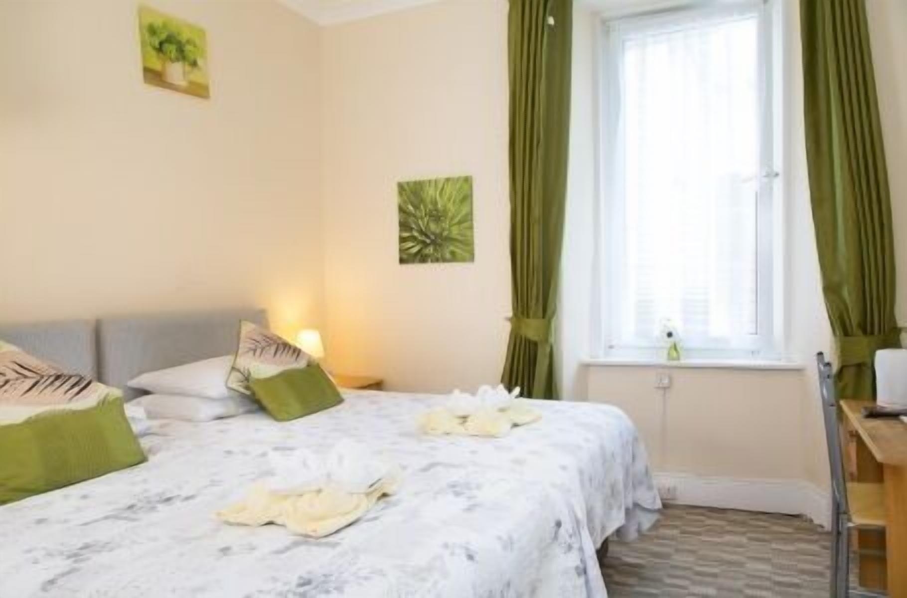 A hotel room with a bed, a nightstand, a window with curtains, and a wall with a painting.